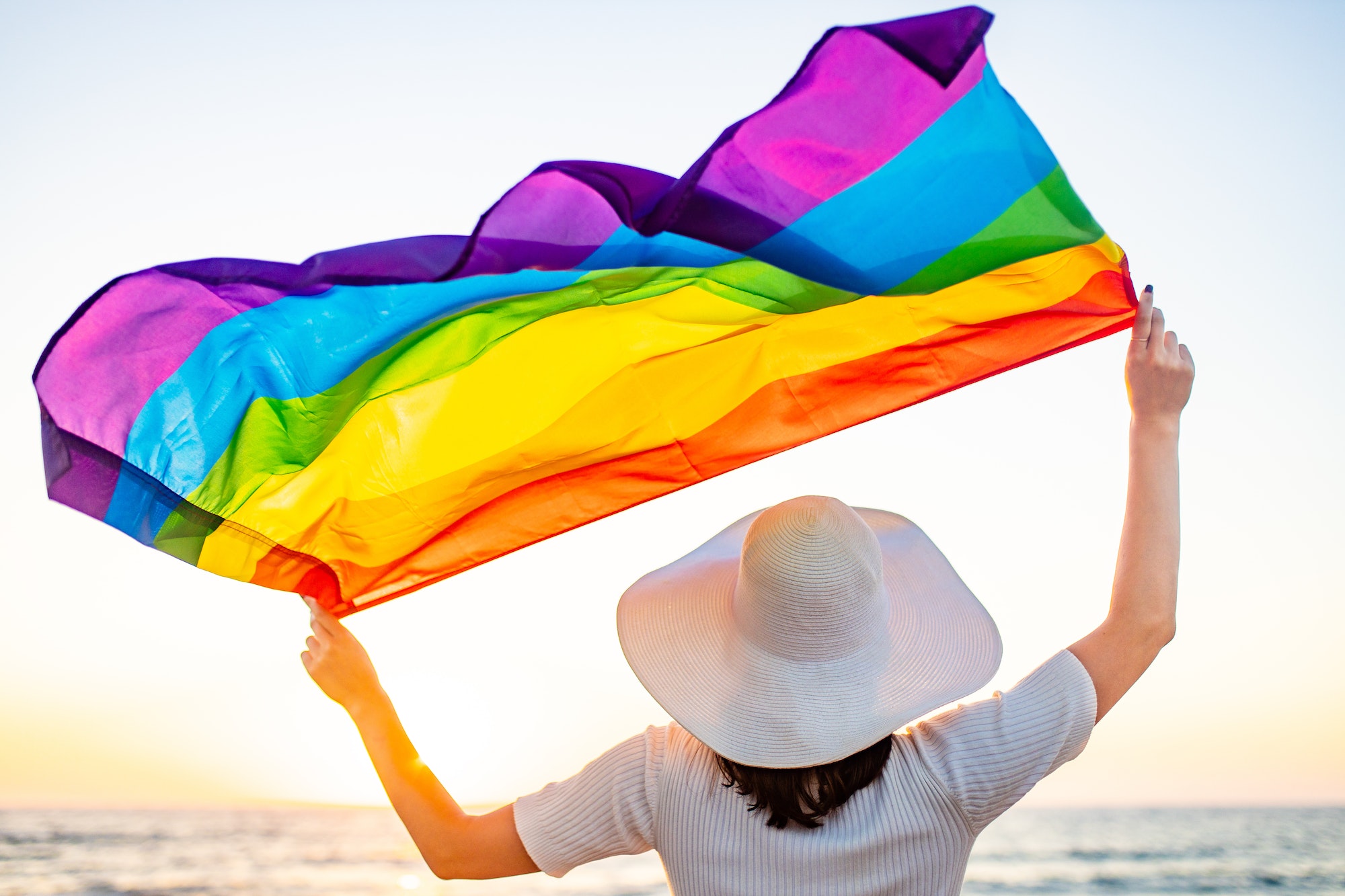 Back view of woman in white dress and hat holding gay pride flag at sunset in beach