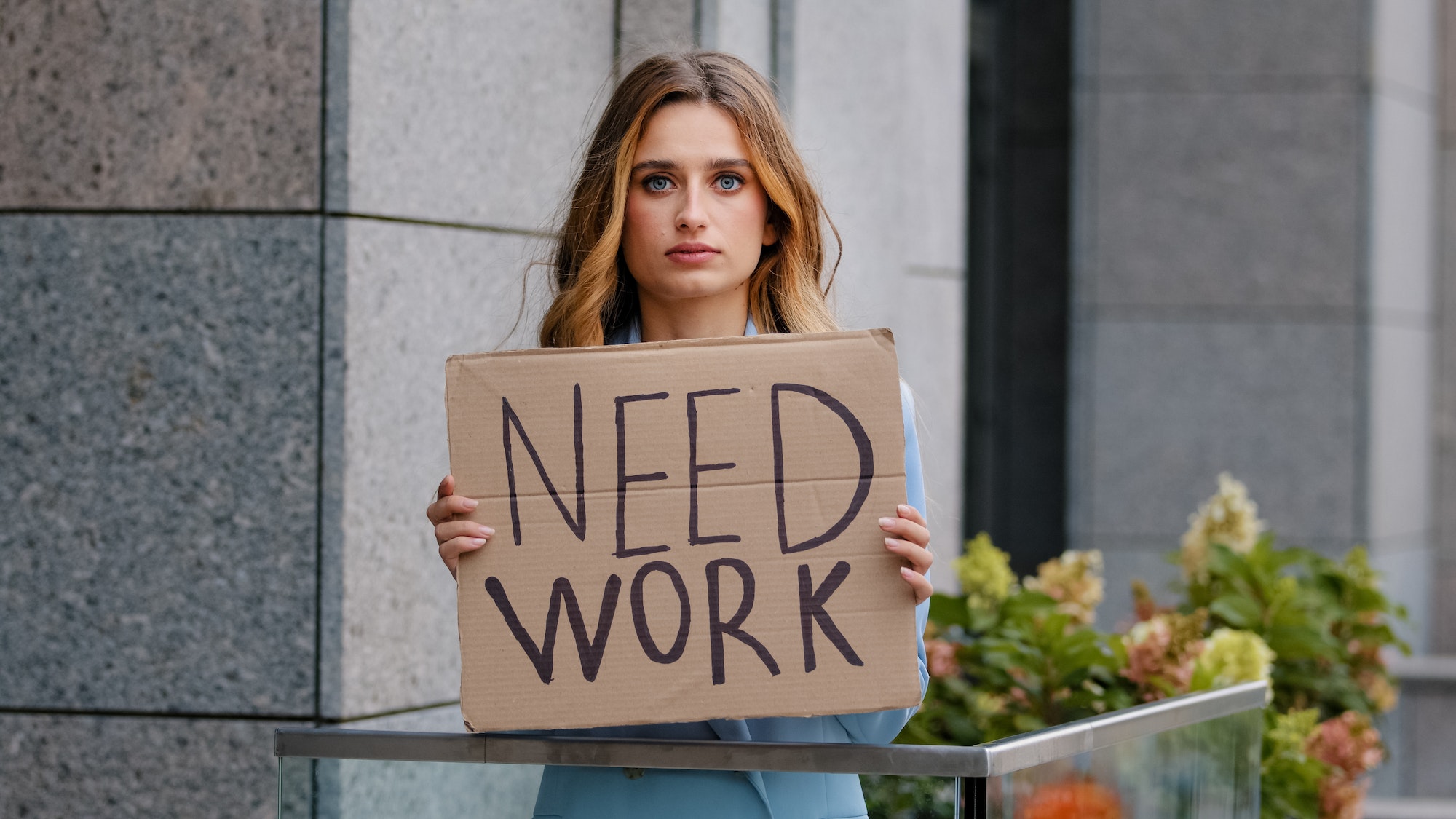 Sad unemployed blonde business woman stands in city looking at camera holds cardboard banner needs